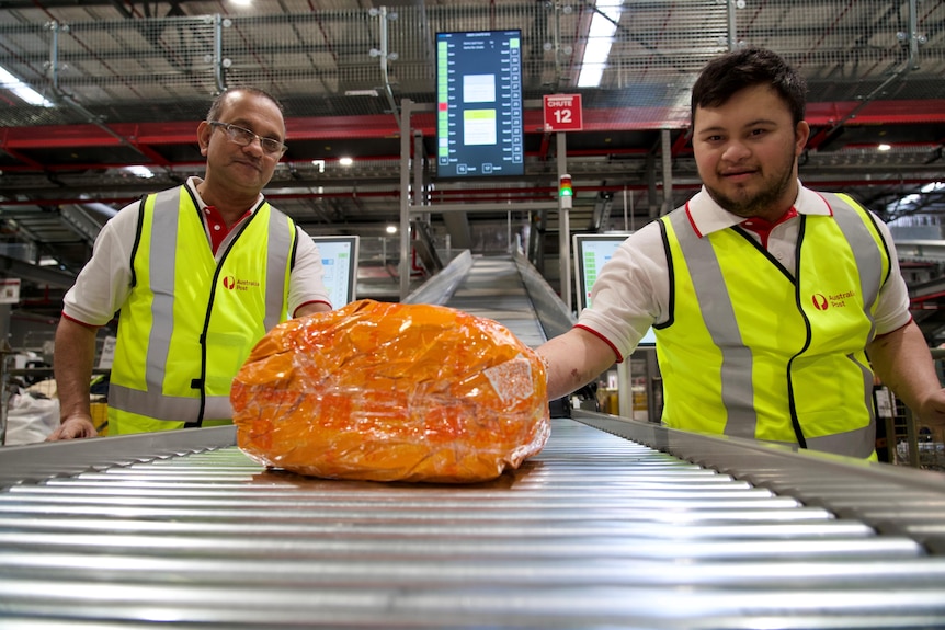 A son and his father in a parcel sorting warehouse standing next to an orange package on a conveyor belt.