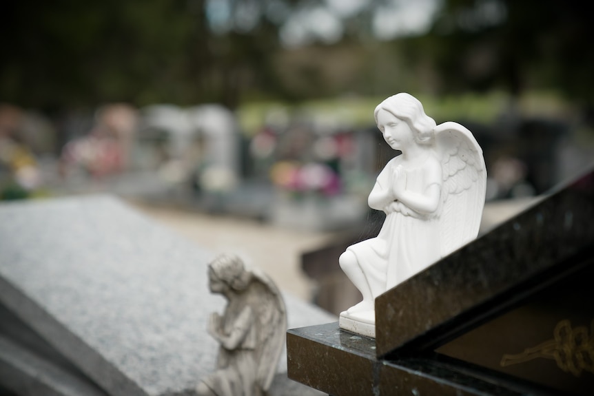 A white statue of an angle atop of a grave stone, amid many other grave stones.
