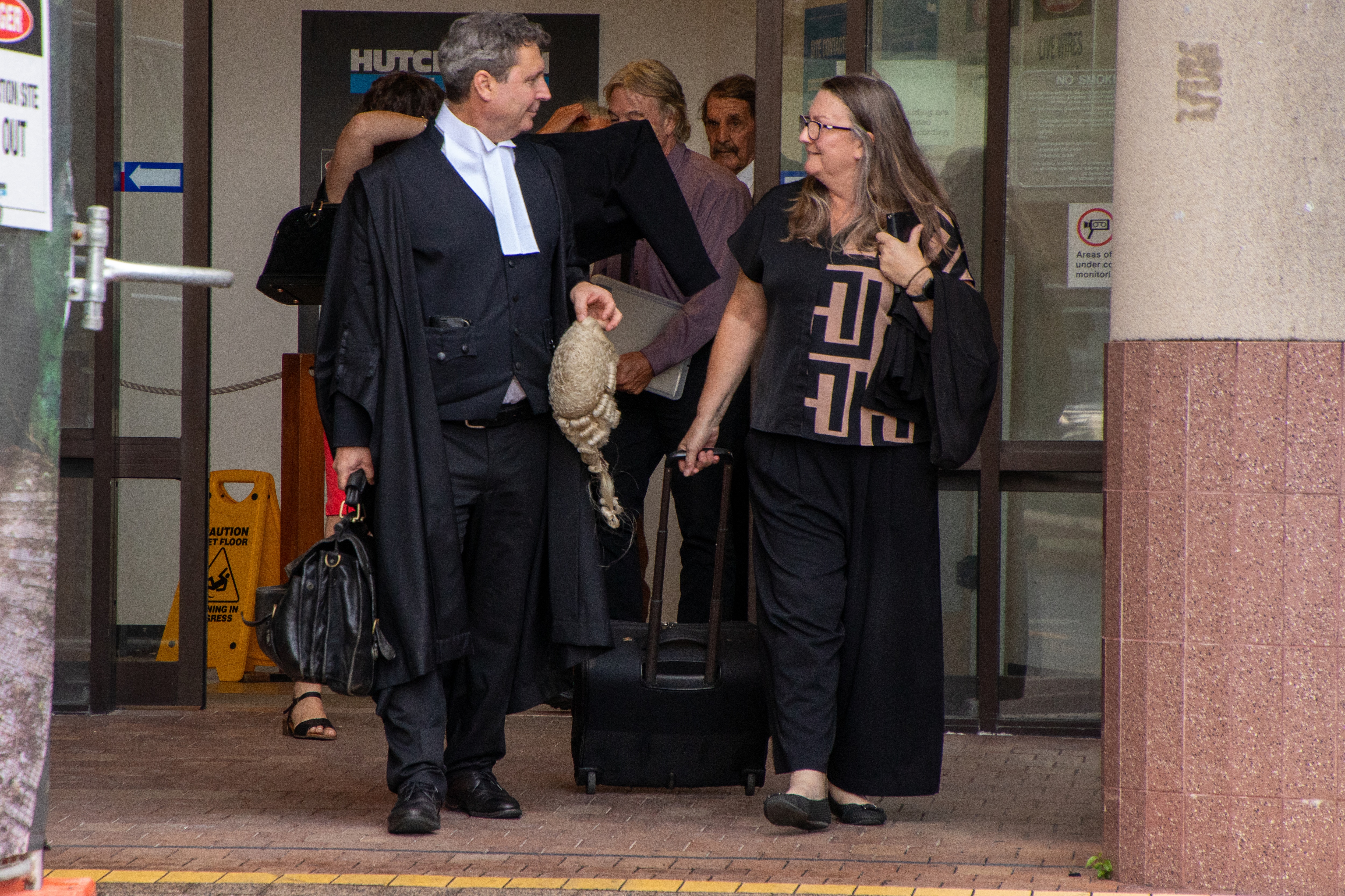 Un hombre de cabello gris vestido con sedas de abogado sostiene una peluca mientras sale de un tribunal al paso de una mujer vestida de oscuro.