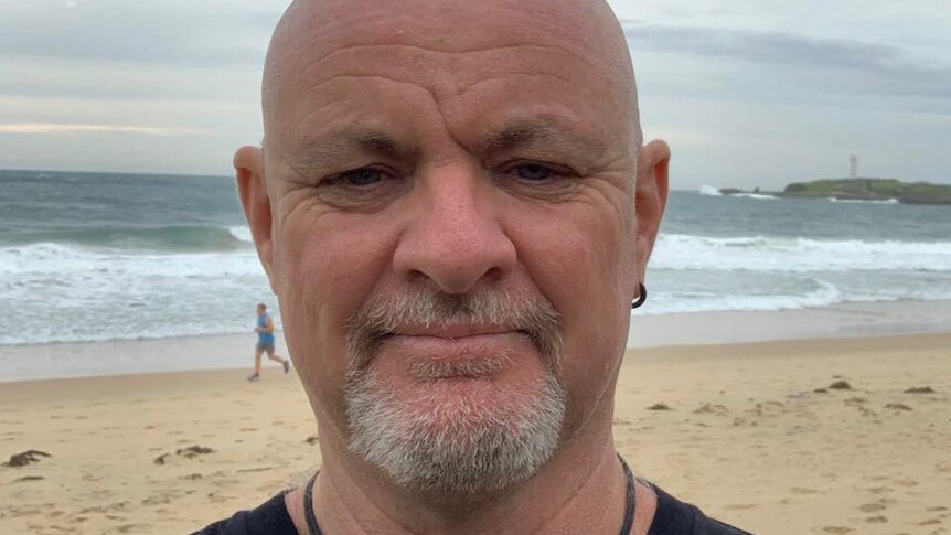 A headshot of Paul Miners, who has a goatee and ear piercing, outside with ocean in the background.