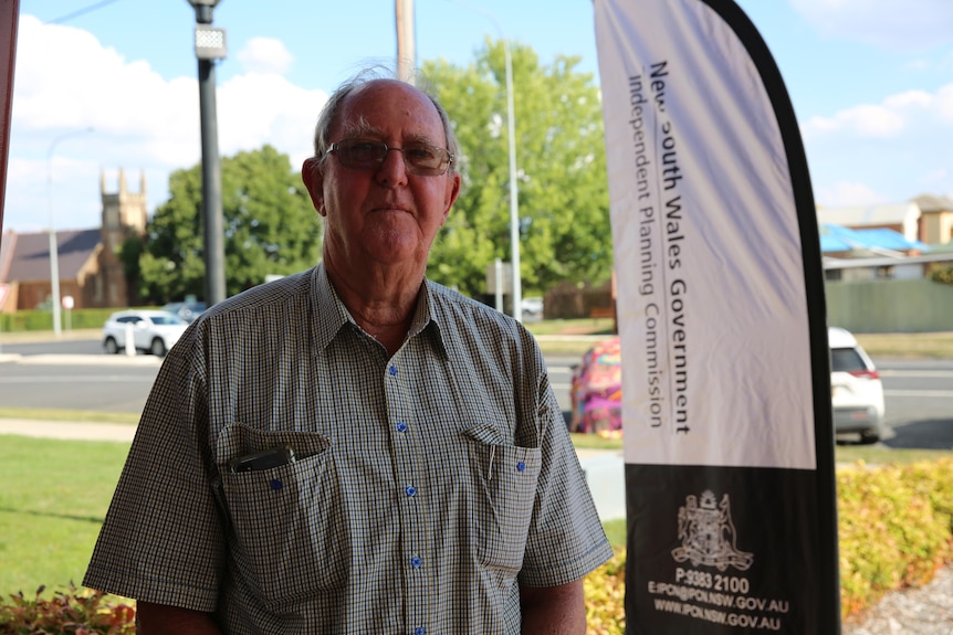 Ajnm older man with balding hair, wearing glasses and a red checked shirt, in front of a NSW government sign