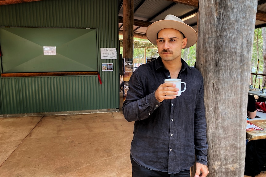 A man with a moustache and nose ring wearing a hat leans against a tree trunk while holding a cup.