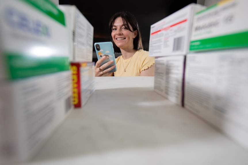 A woman wearing a yellow top looks at her phone with some boxes of medication in front of her