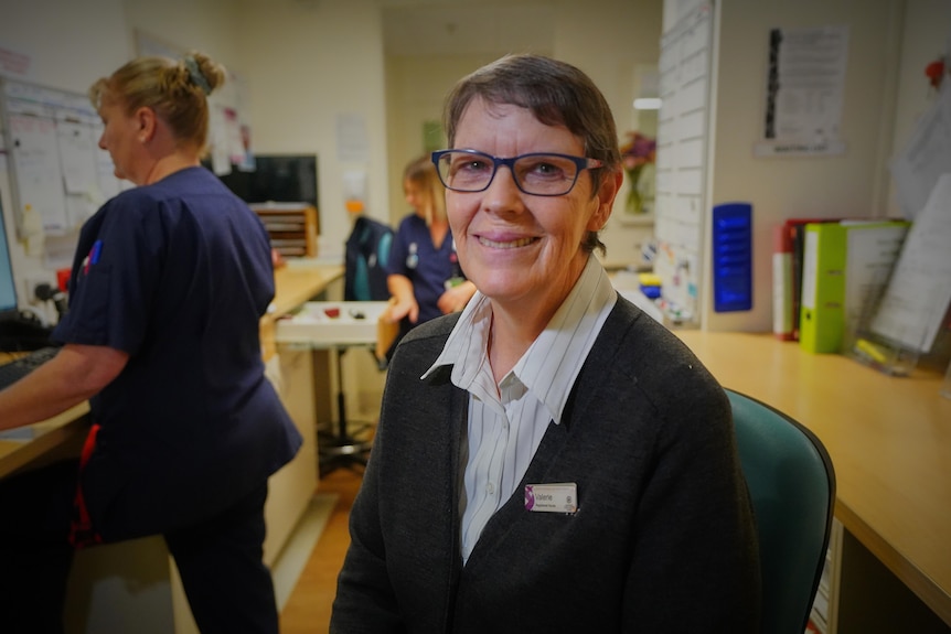 A woman sitting behind a nurses' station smiles at the camera