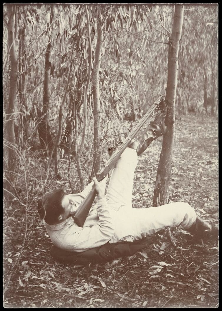 a man lies on the ground with his leg brace on a tree, nestling a rifle that he's aiming up into the canopy