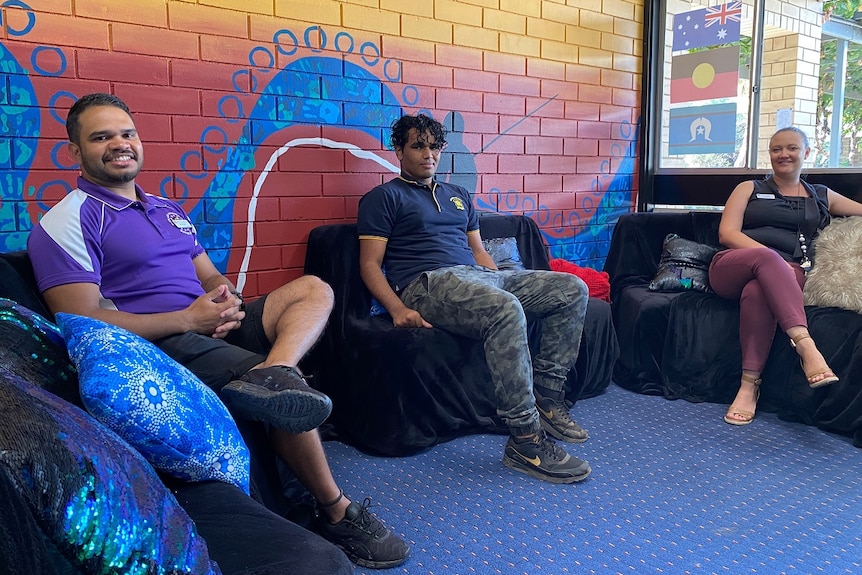 Two men and a woman are sitting on couches, smiling. There's a colourful mural and three flags behind them.