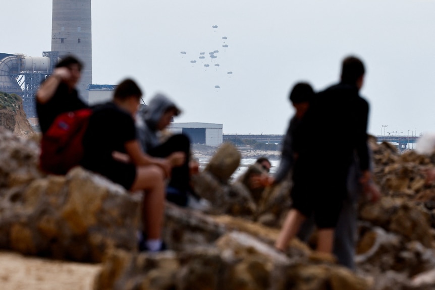 Aid falls to the ground attached to parachutes in the distance as a group of people sit on rocks looking on