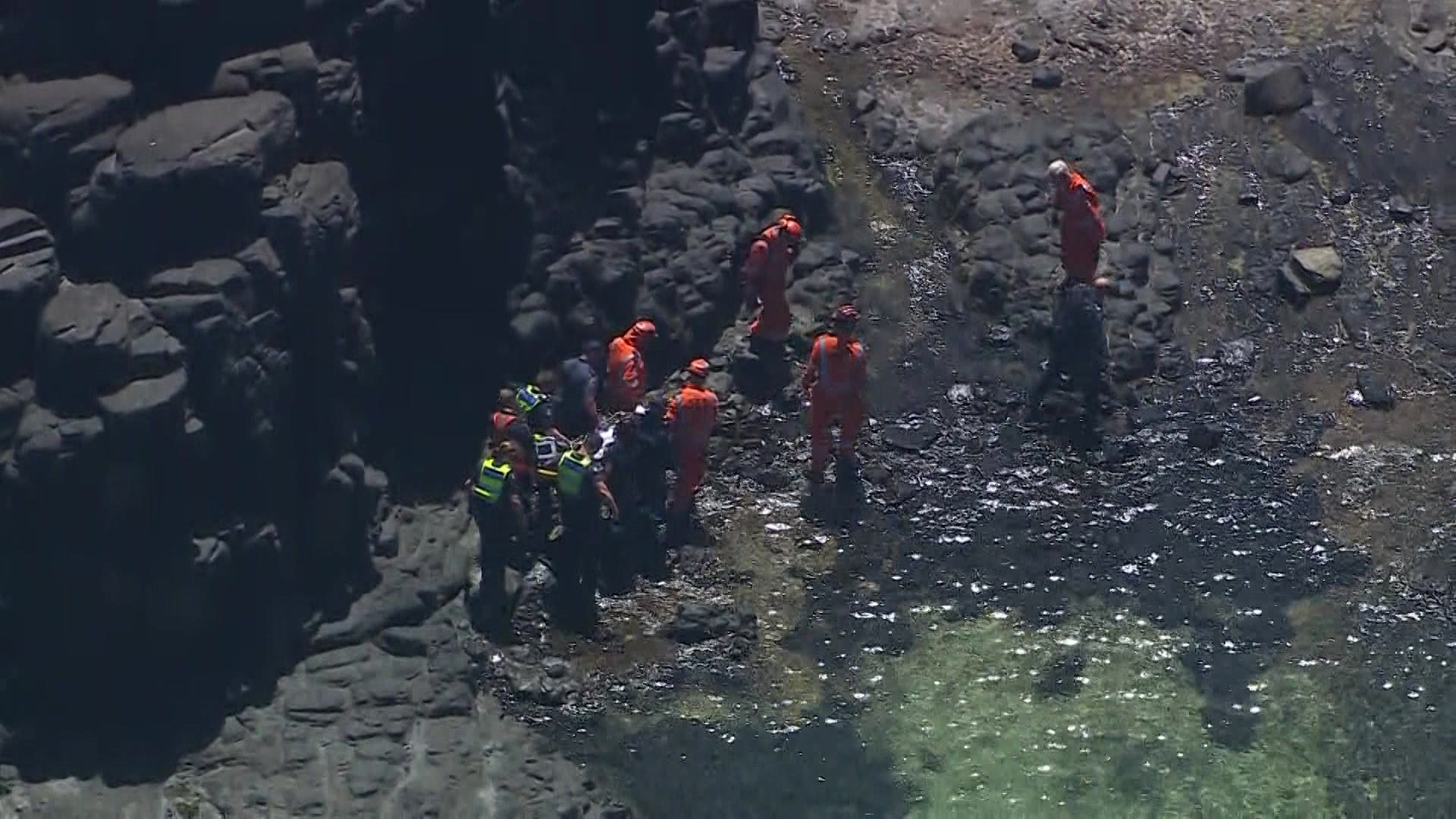 Personas con monos azul marino y naranja caminan sobre rocas a través del agua con una camilla en la mano.