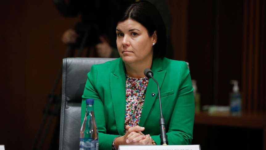 A woman in a green blazer sits behind a desk with a microphone