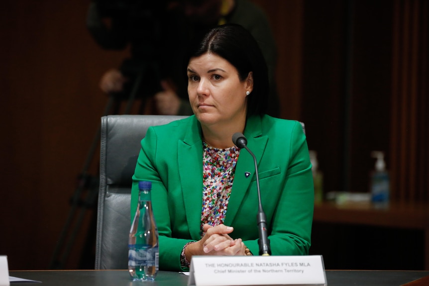 A woman in a green blazer sits behind a desk with a microphone