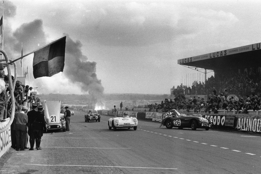 In this black and white photo, three cars are on the main straight at Le Mans, while a massive fire ball is in the background.
