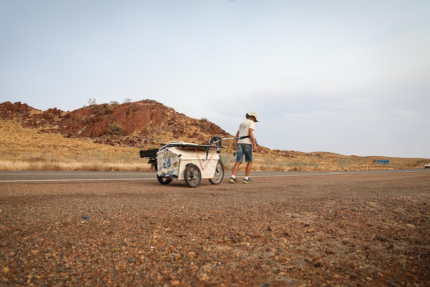An older man tows his small cart as he walks along a highway surrounded by red hills. 