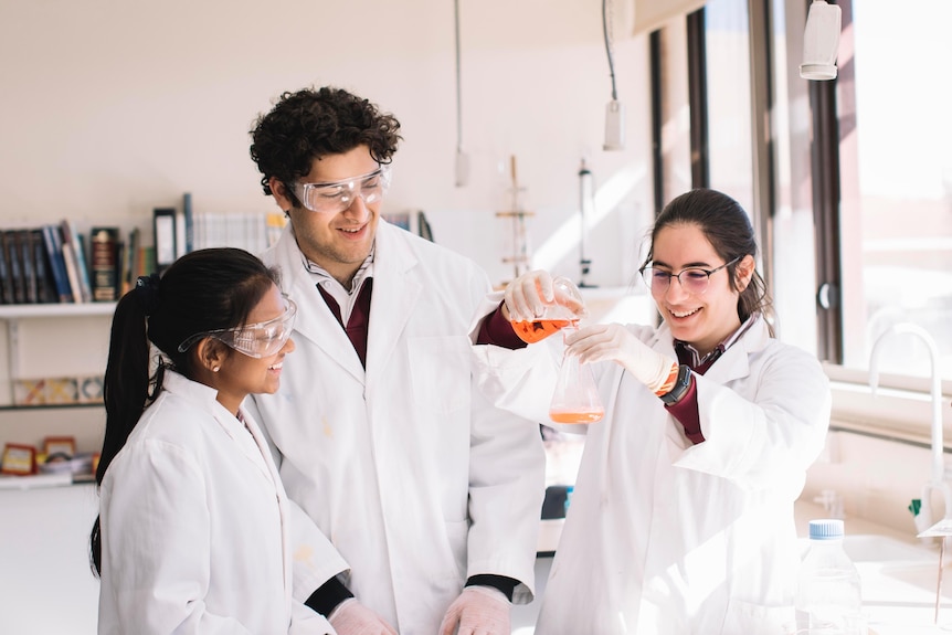 Three students in white lab coats look at a beaker full of liquid.