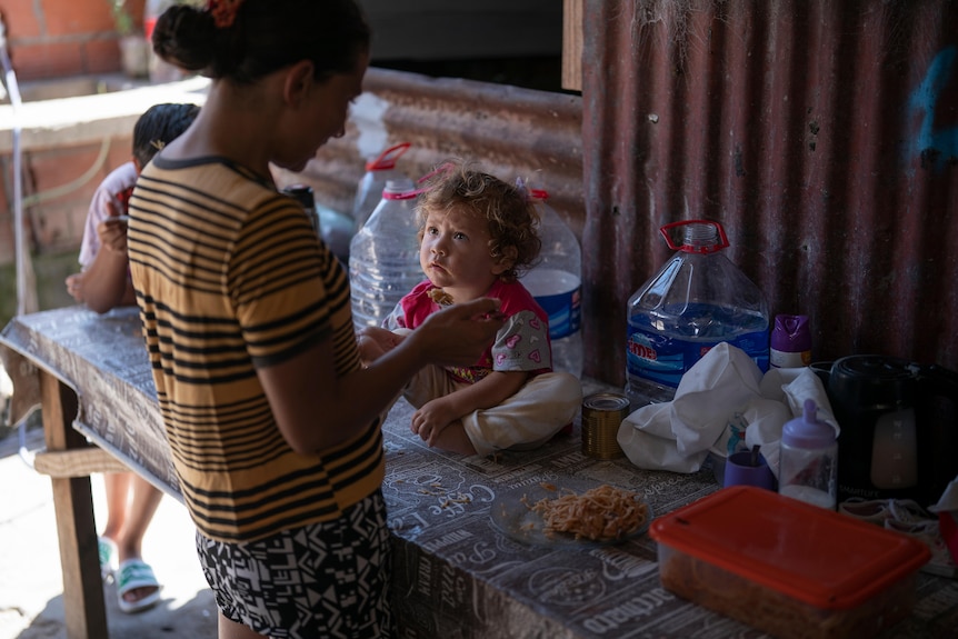 A young woman feeds a small child sitting on a table amongst some plastic containers, in a shelter made of corrugated iron.