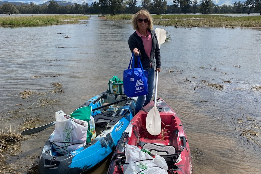 A woman stands up in a kayak holding onto a paddle in one hand and a bag of groceries in the other.