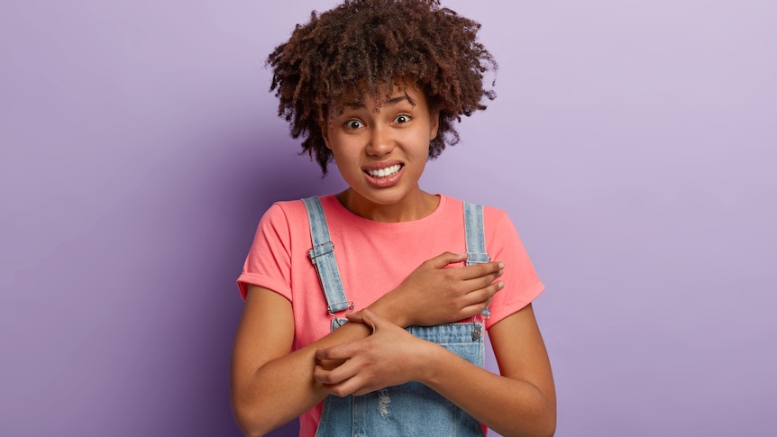 A displeased woman wearing a pick t-shirt and blue overalls scratching an itch on her arm