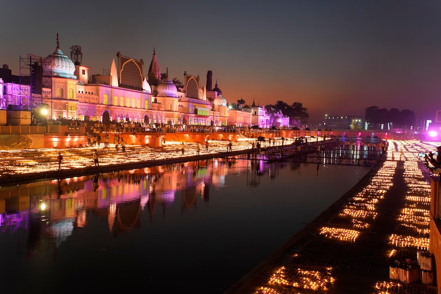 Thousands of small lanterns lit along a riverbank as Indian buildings are illuminated by colourful lights. 