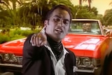 Pauly Fuemana wearing a black jacket and cravat points to the camera with a red convertible sports car behind him.