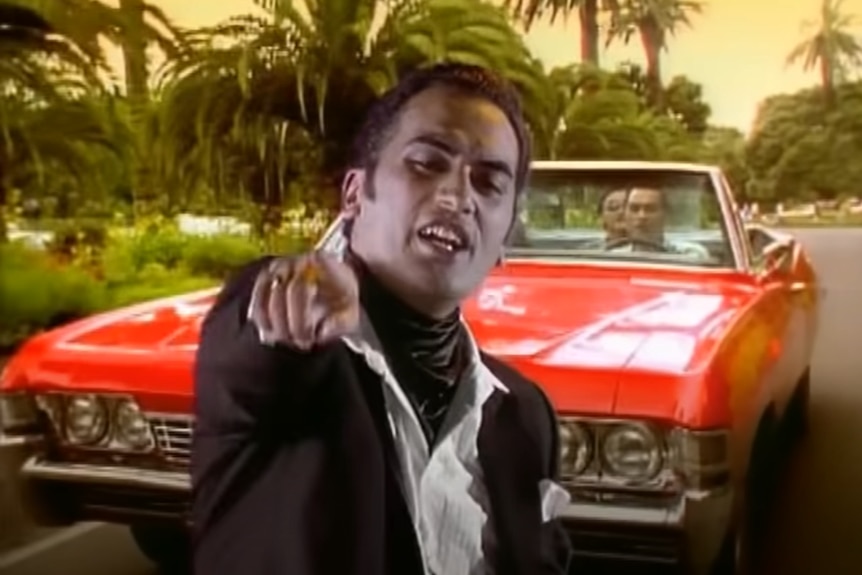 Pauly Fuemana wearing a black jacket and cravat points to the camera with a red convertible sports car behind him.