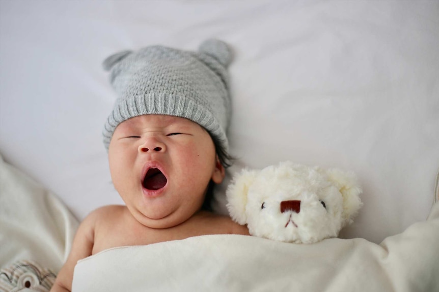 Cute yawning baby wearing a grey beanie in a bed for a story about why parents are choosing masculine names for their daughters.