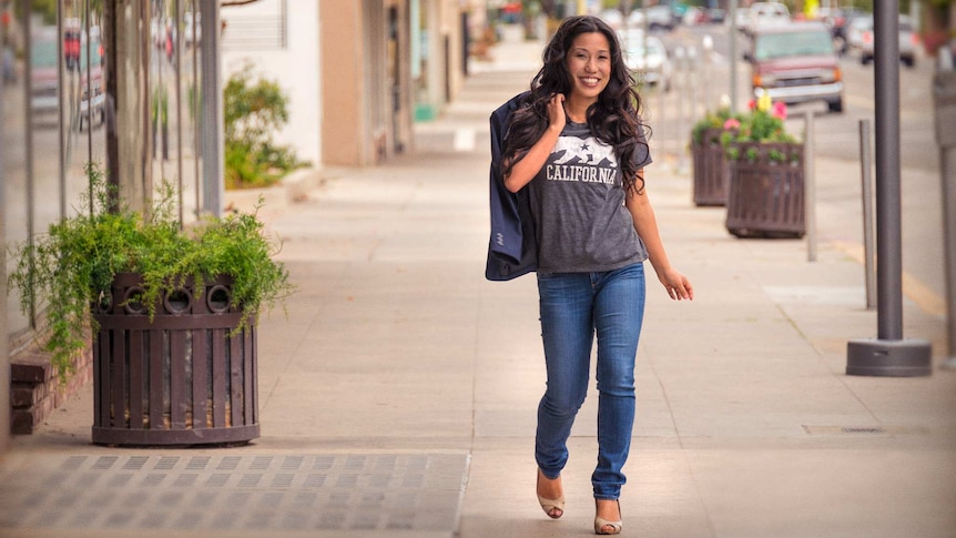 A portrait of Elizabeth Heng walking down a high street in jeans, heels and a t-shirt that reads 'California'.