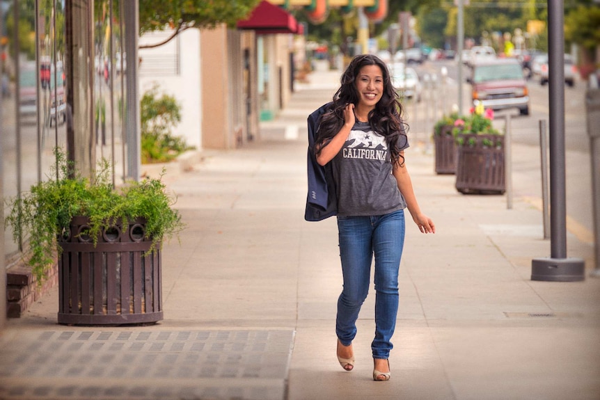 A portrait of Elizabeth Heng walking down a high street in jeans, heels and a t-shirt that reads 'California'.