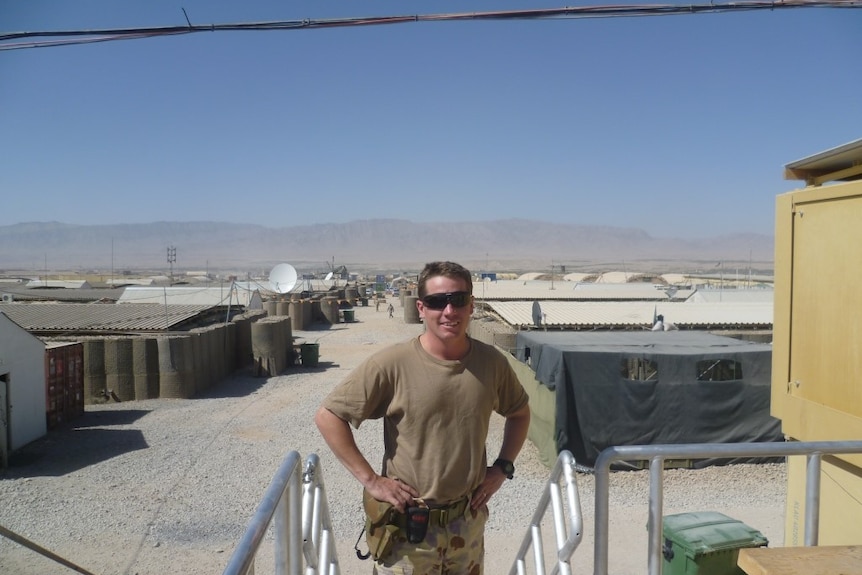 Man standing in desert army fatigues wearing black sunglasses with hands on hips. Dry desert dusty conditions.