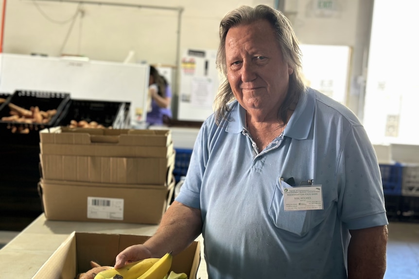 a man with grey hair is slightly smiling. he is wearing a blue polo shirt and is in a warehouse with boxes of groceries