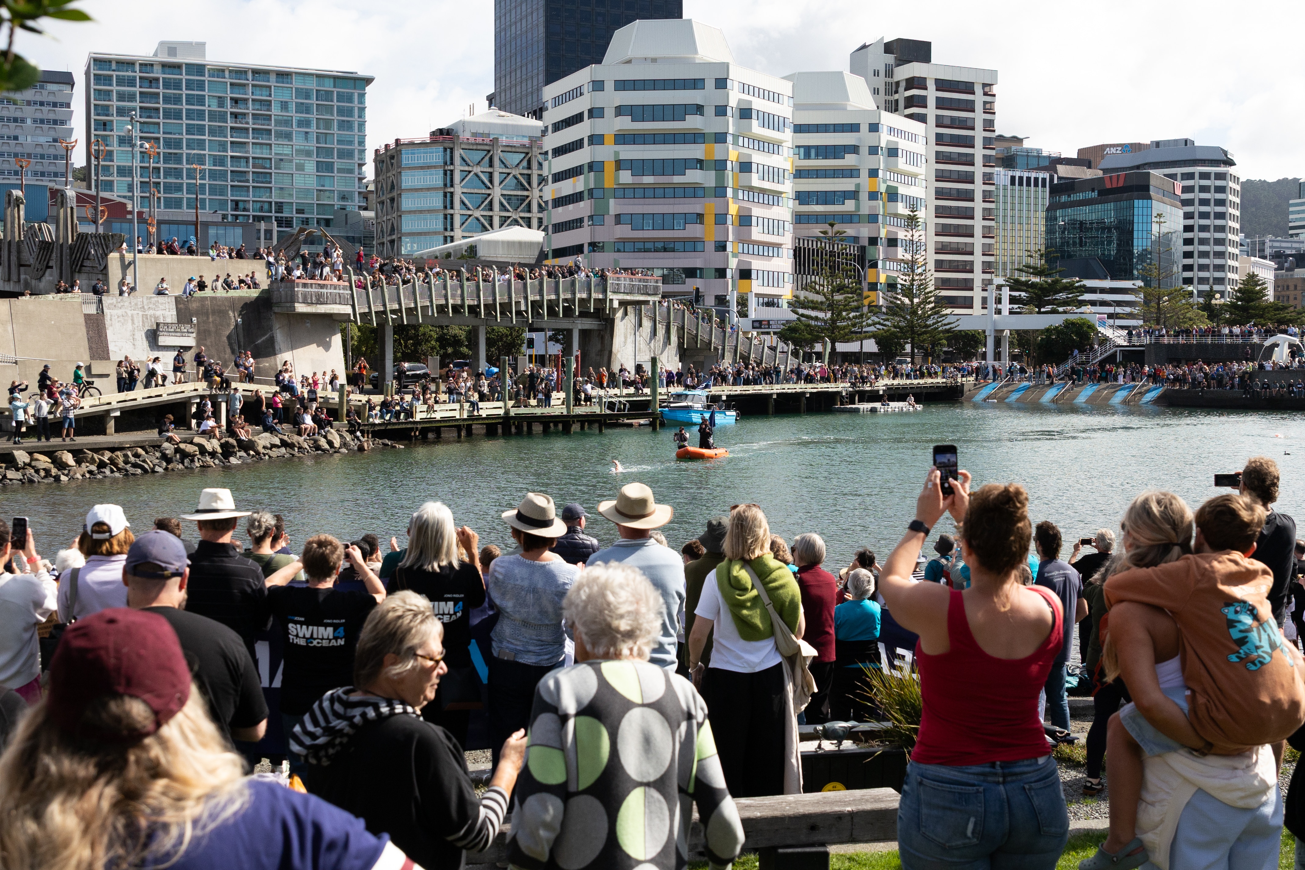 Crowds of people line harbours in a city, watching the water.