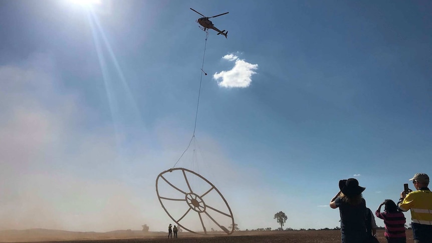 Several people gather watching a helicopter taking off, hoisting with it a huge circular device hanging below.