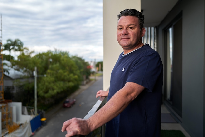 A man standing on a balcony, with his hands on the railing, turns to look at the camera with a neutral expression.