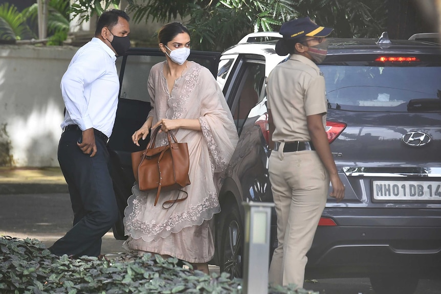 A woman in floaty outfit steps out of a car wearing a mask. An official in uniform stands nearby.