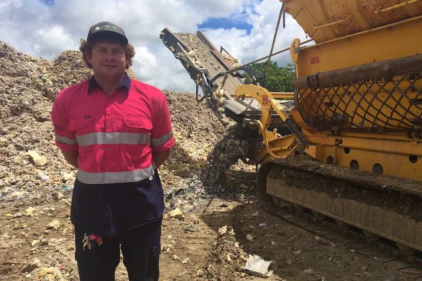 Man stands next to a giant shredder on his right. behind him is a pile of shredded mattresses and pile of steel