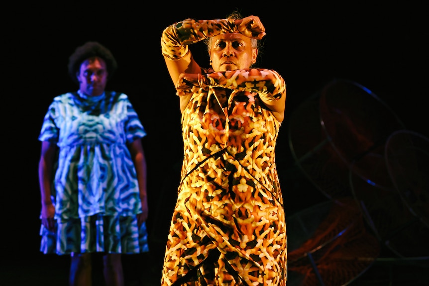 A person in a brightly patterned outfit holds their arms out in front of their face on stage.