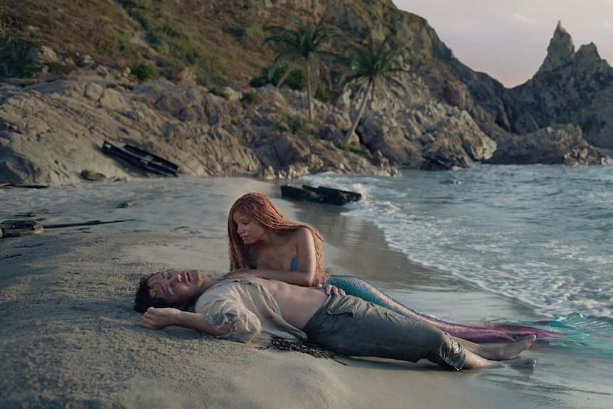 A young black mermaid with a green tail lays beside a man who has washed up on the shores of a beach, and seems unconscious.