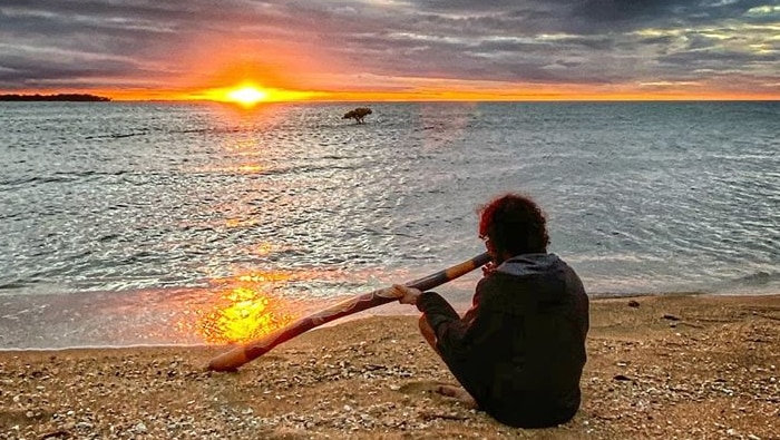 Butchulla man Dinka Dinka  sits on the beach holding a didgeridoo at sunset