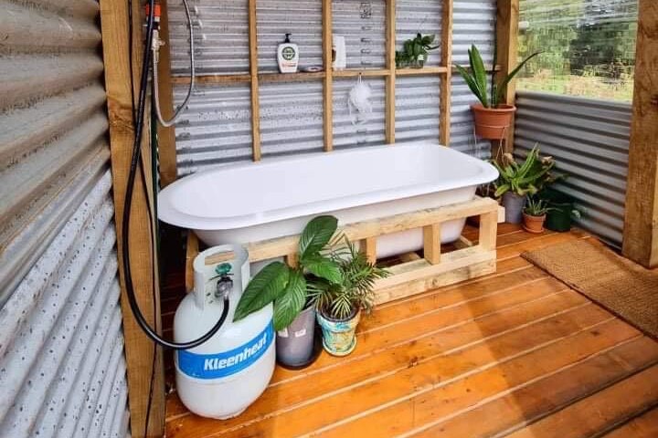 An outdoor bathroom inside a tin shed-like building. A white bathtub, shower and plants with a simple gas heater