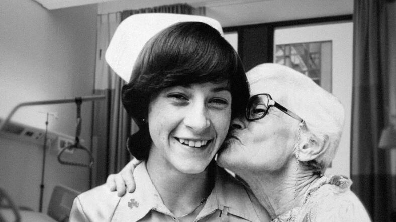 A woman with dark hair wearing a nurses uniform smiles as she is kissed on the cheek by an elderly woman in a hospital room.