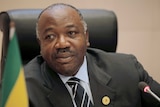 Gabon's President Ali Bongo Ondimba looks at the camera as he sits next to a Gabon flag.