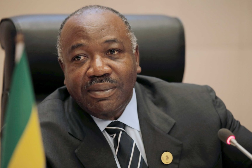Gabon's President Ali Bongo Ondimba looks at the camera as he sits next to a Gabon flag.