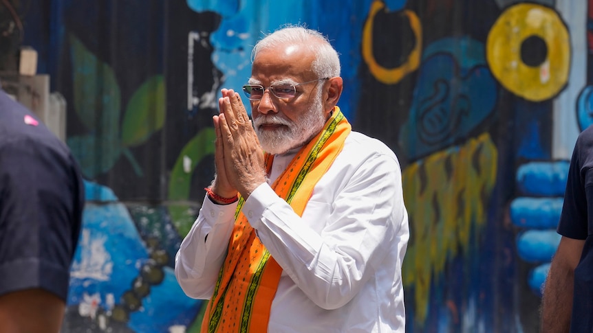 Indian prime minister Narendra Modi, wearing a white shirt and orange scarf, holds his hands together in prayer