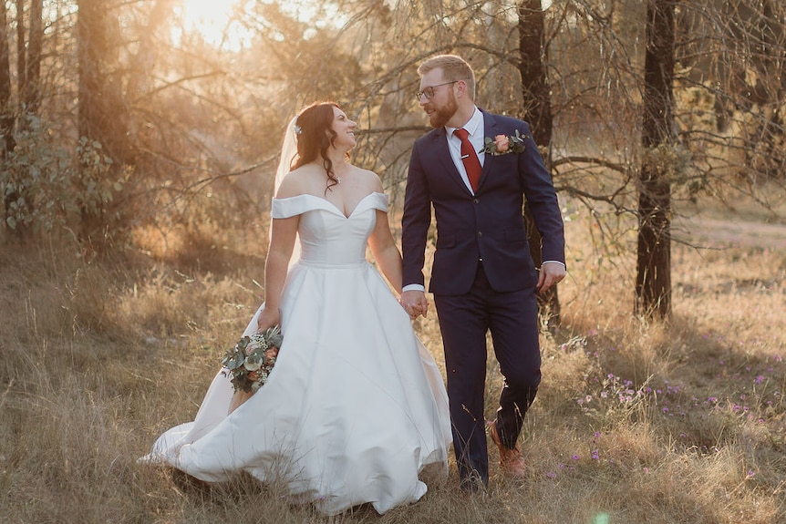 Una novia sonriente y su nuevo marido caminan por un claro de arbustos.