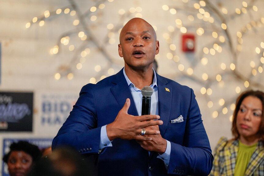 A black bald man holds a microphone while he speaks in front of an array of fairy lights 
