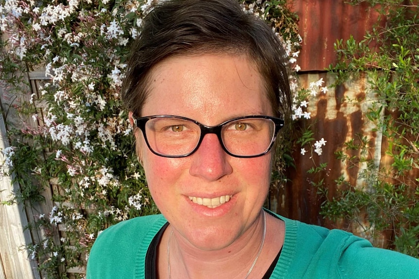 A woman with short dark hair and glasses takes a selfie in front of a garden fence.