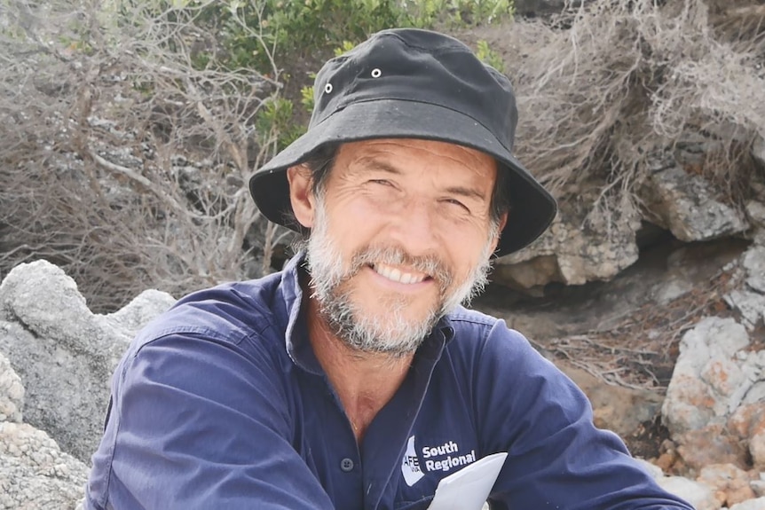 He sits in a bucket hat at the beach, smiling at the camera