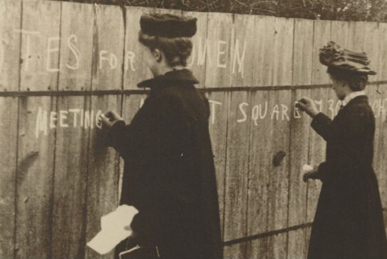 Three women writing pro-suffragette graffiti on a wall in chalk between 1900 and 1910.
