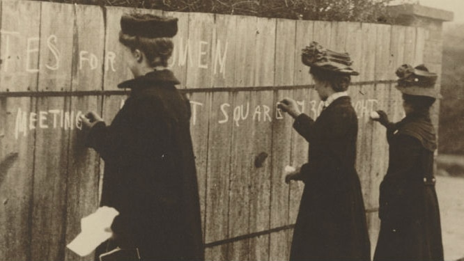 Three women writing pro-suffragette graffiti on a wall in chalk between 1900 and 1910.