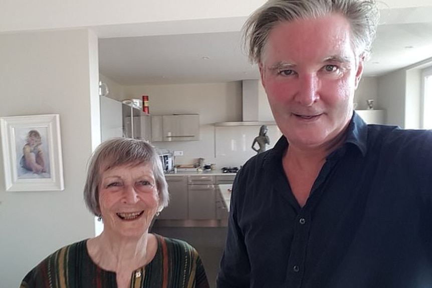In a kitchen, an elderly woman with wide smile and grey hair stands near a younger man, with more subtle smile. Both face camera