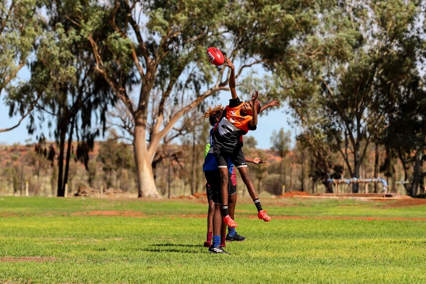 Two young Aboriginal women wearing football jumpers jump at a football thrown by an umpire
