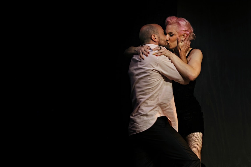 A 40-something bald man wearing an untucked shirt and a pink-haired 30-something trans woman kiss passionately 